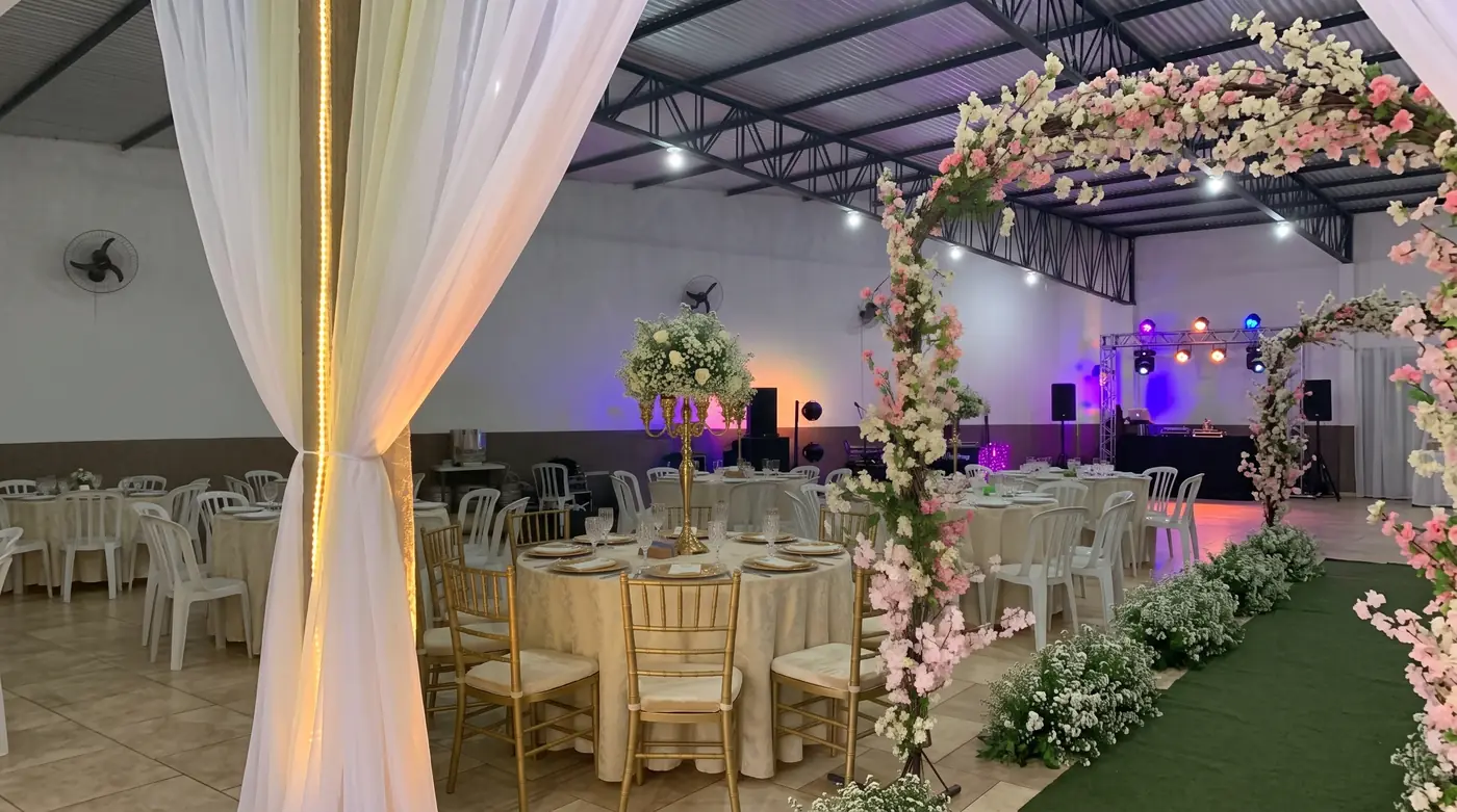 Salão decorado para casamento com arco de flores, mesas redondas e cadeiras douradas